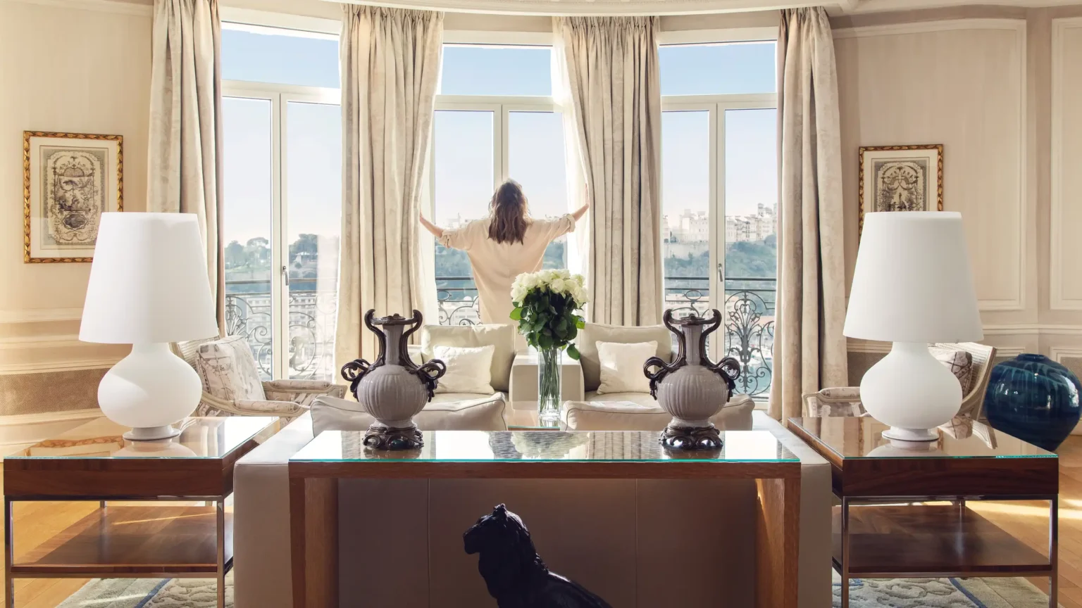 kobieta otwierająca okna w salonie hotelowym, ubrana w szlafrok i odwrócona tyłem do obiektywu