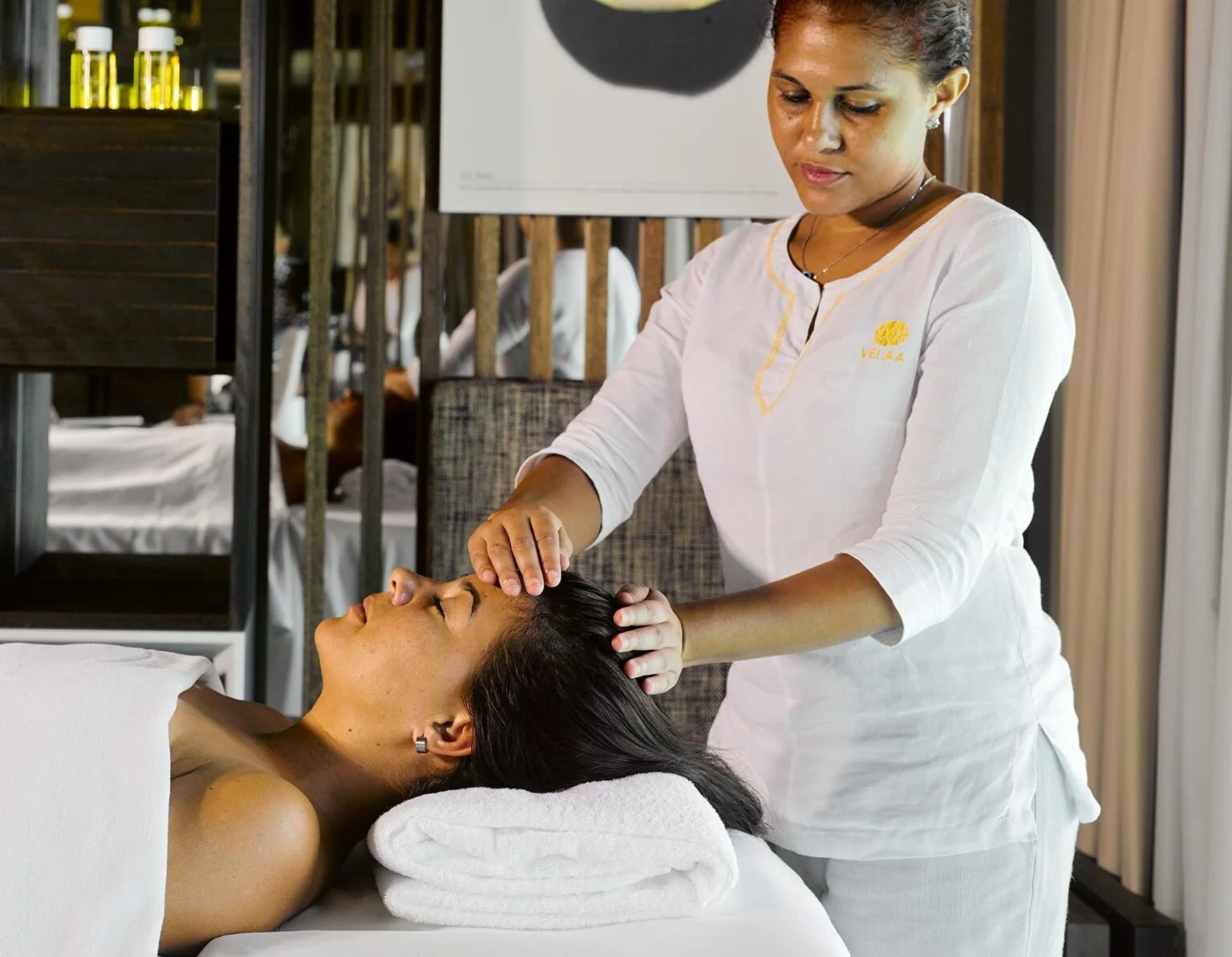 a dark-skinned woman in a white apron gives a massage to another woman who lies on her back under a white towel