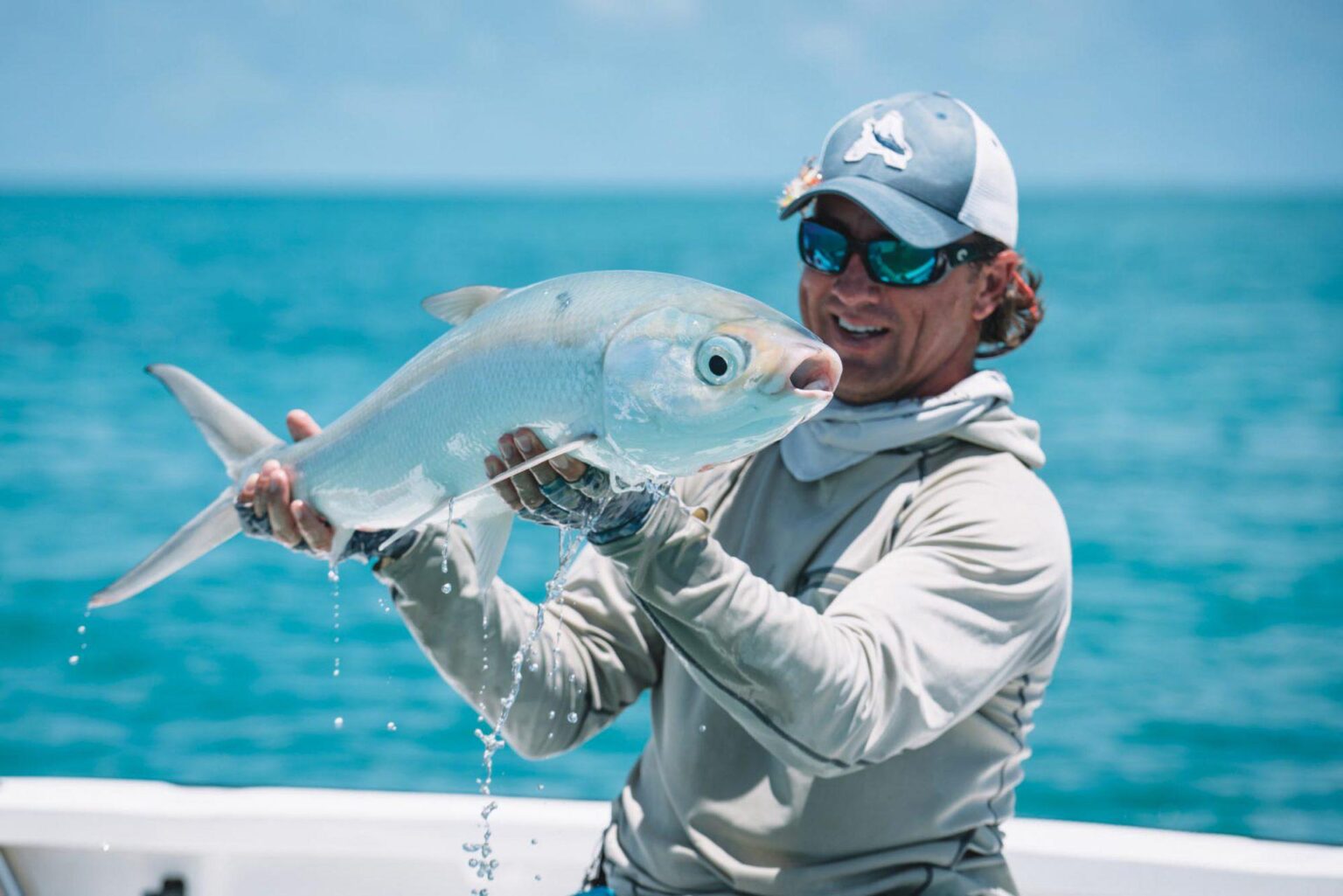 mężczyzna w białej bluzie i czapce trzyma białą rybę na tle oceanu