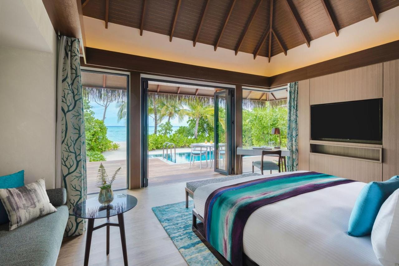 pullman maldives all inclusive resort przykładowa sypialnia z widokiem na otaczającą roślinność i ocean, wyoskie sklepienie dachu w brązowym kolorze, jasna i przestronna sypialnia