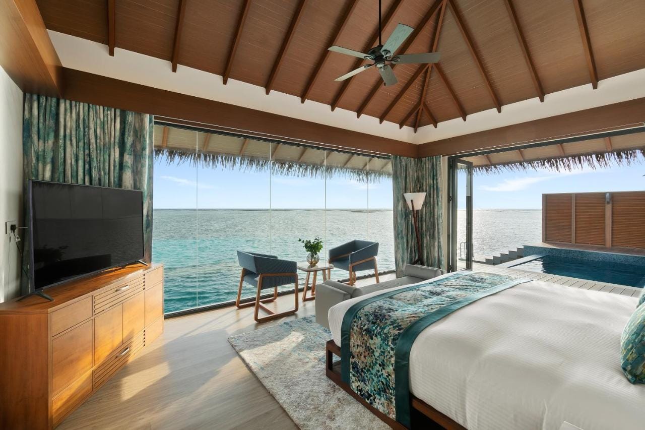 pullman maldives all inclusive resort przykładowa sypialnia z widokiem na otaczającą roślinność i ocean, wyoskie sklepienie dachu w brązowym kolorze, jasna i przestronna sypialnia